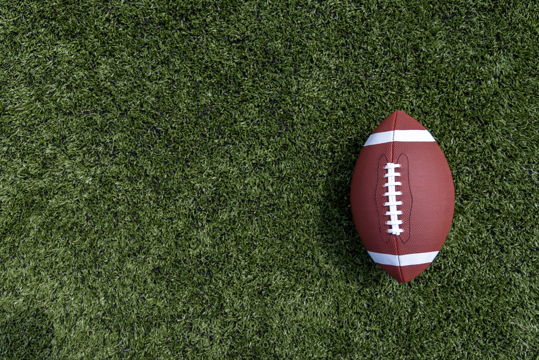 A brown football with white laces lying on green turf field.